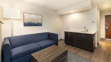 Una sala moderna con un sofá azul, una mesa de centro de madera, una zona de cocina con armarios oscuros y un cuadro en la pared.