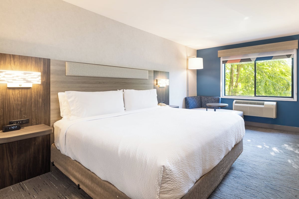 A hotel room with a large bed, bedside tables, a desk, and a chair. There is a window with a view of trees.