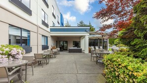 An outdoor patio area with seating and a covered section, surrounded by greenery and a building with large windows.