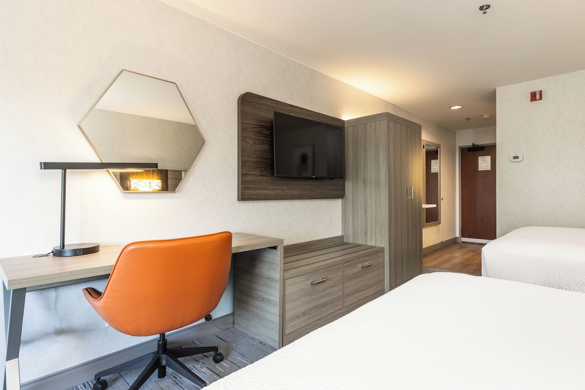 A hotel room with a desk, an orange chair, a bed, a TV, and a painting on the wall.