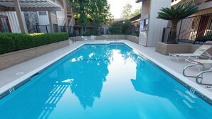 A rectangular swimming pool with clear blue water, surrounded by a low wall and a paved deck. There are lounge chairs and a small table on the deck.