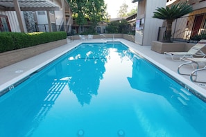 A rectangular swimming pool with clear blue water, surrounded by a low wall and a paved deck. There are lounge chairs and a small table on the deck.