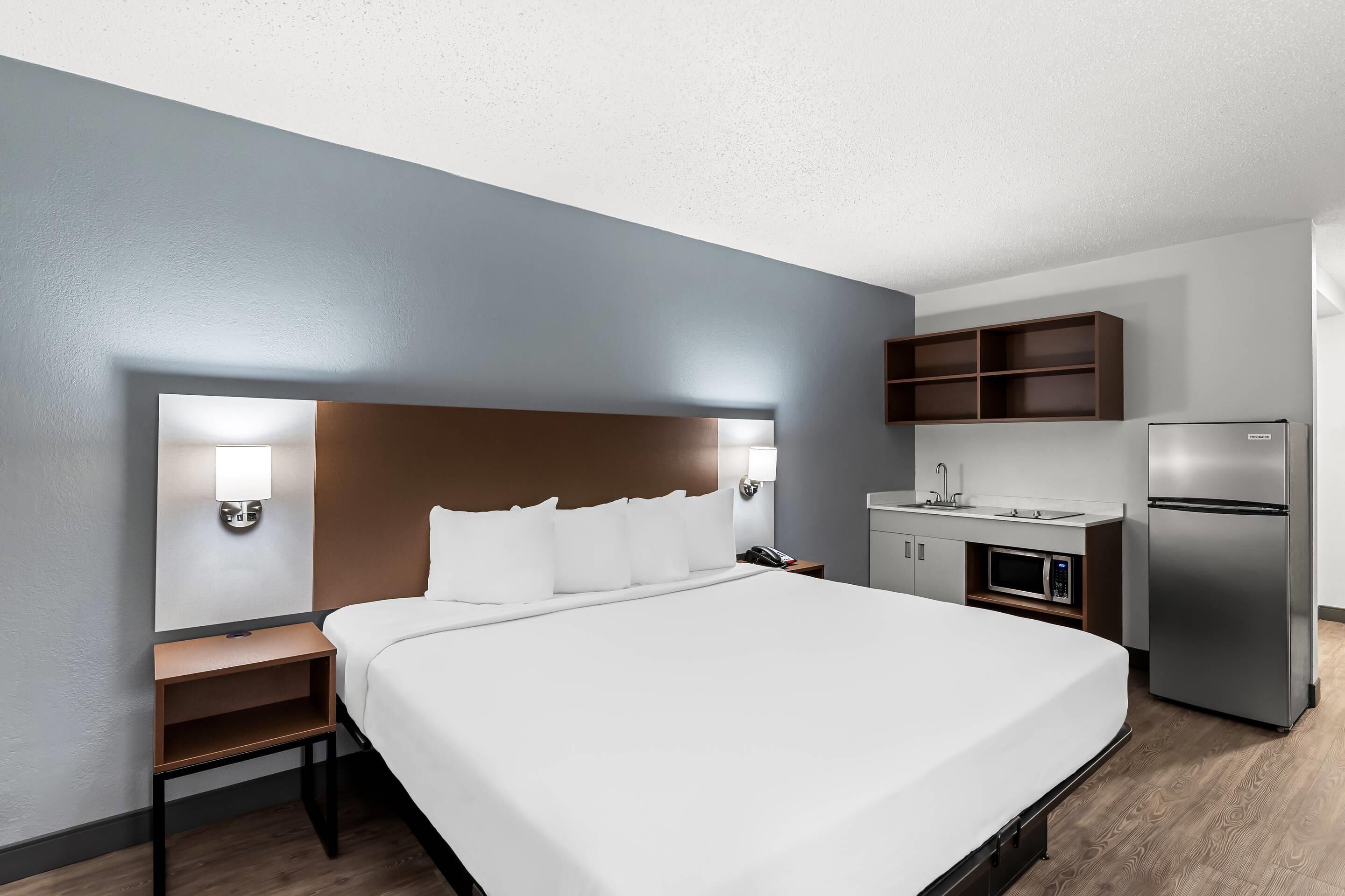 A hotel room with a bed, bedside table, lamp, and a kitchenette area including a microwave and refrigerator.
