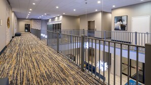 A hotel corridor with a patterned carpet, a railing, and a reception desk with a glass partition. - Baymont by Wyndham White Plains - Elmsford (Elmsford)