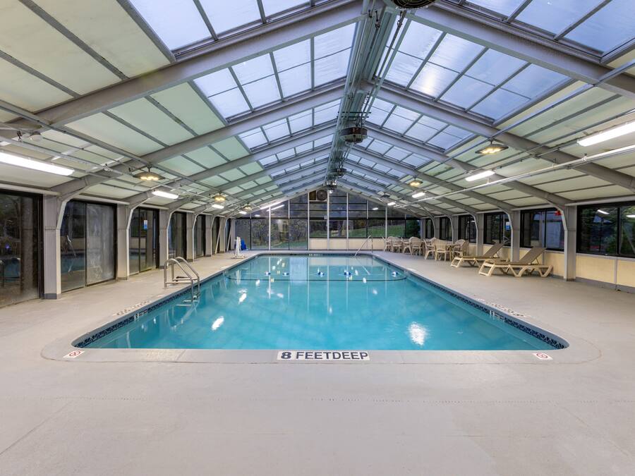 An indoor swimming pool with a glass ceiling, clear signage indicating depth, and lounge chairs along the sides.
