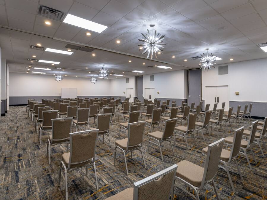 Un salón de conferencias amplio con filas de sillas, luces en el techo y una pizarra.