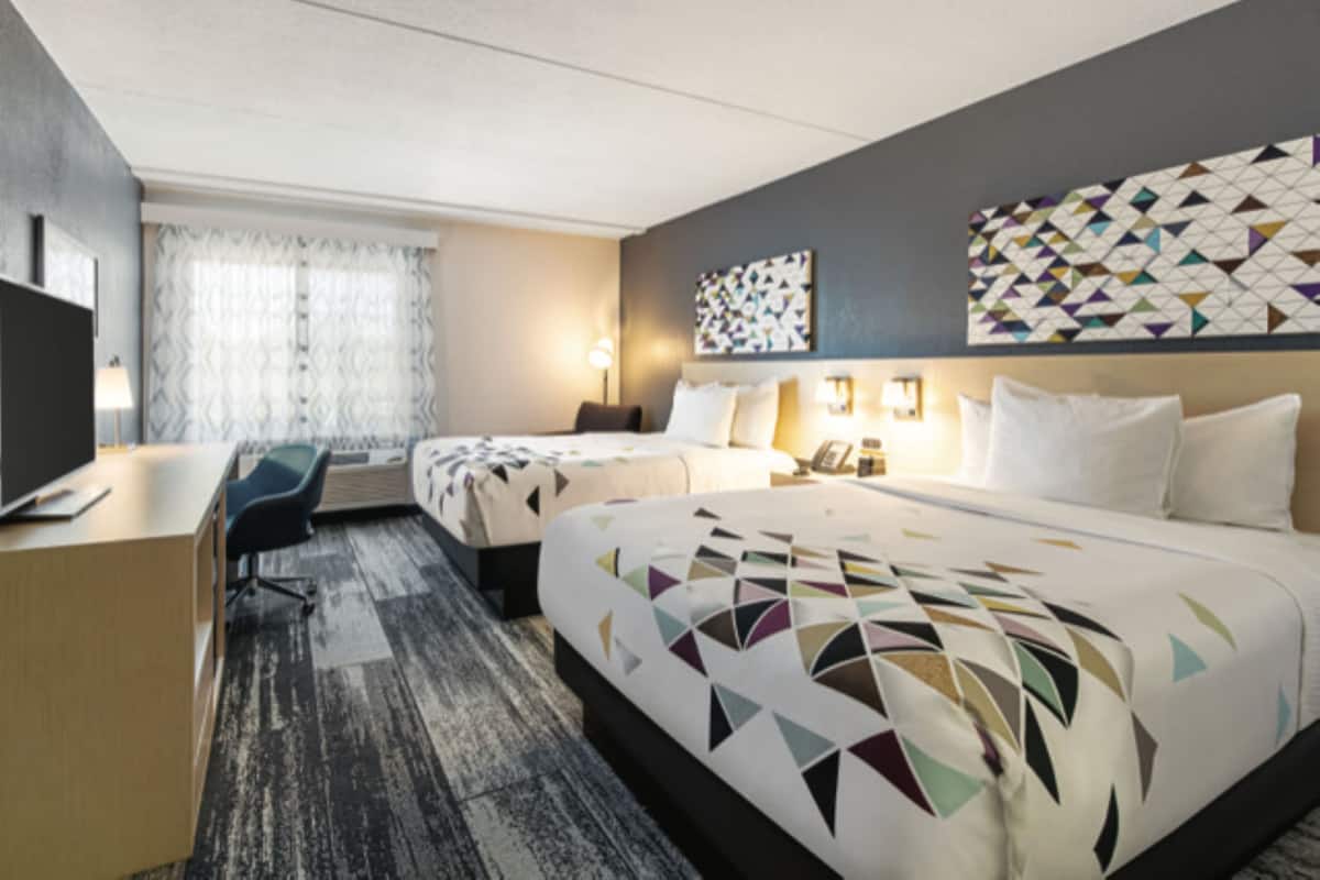 Ein Hotelzimmer mit einem großen Bett, einem Schreibtisch und einem Stuhl. Ein Fenster mit Vorhängen und eine Wand mit geometrischem Muster.