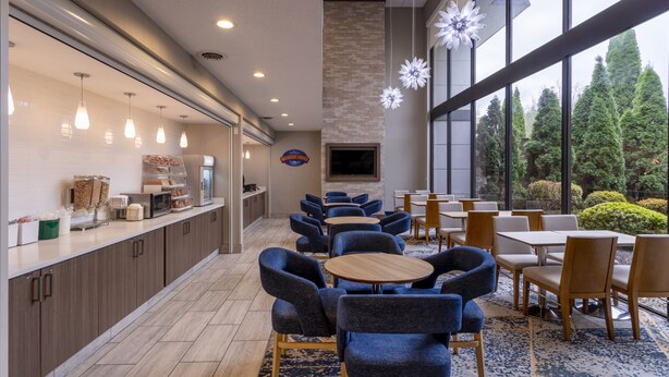 A modern hotel lobby with a kitchen area, seating arrangements, and large windows offering a view of the outdoors. - Baymont by Wyndham White Plains - Elmsford (Elmsford)