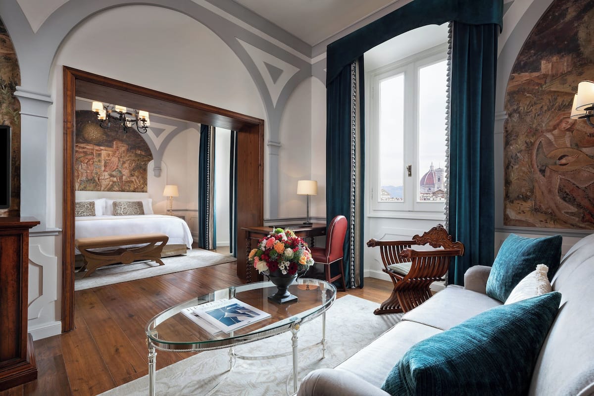 A hotel room with a bed, a sitting area with a sofa and armchair, a glass coffee table with a vase of flowers, and a view of a cityscape through a window.
