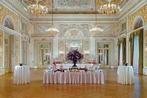 An opulent banquet hall with ornate chandeliers, marble walls, and a central table set for a formal event.