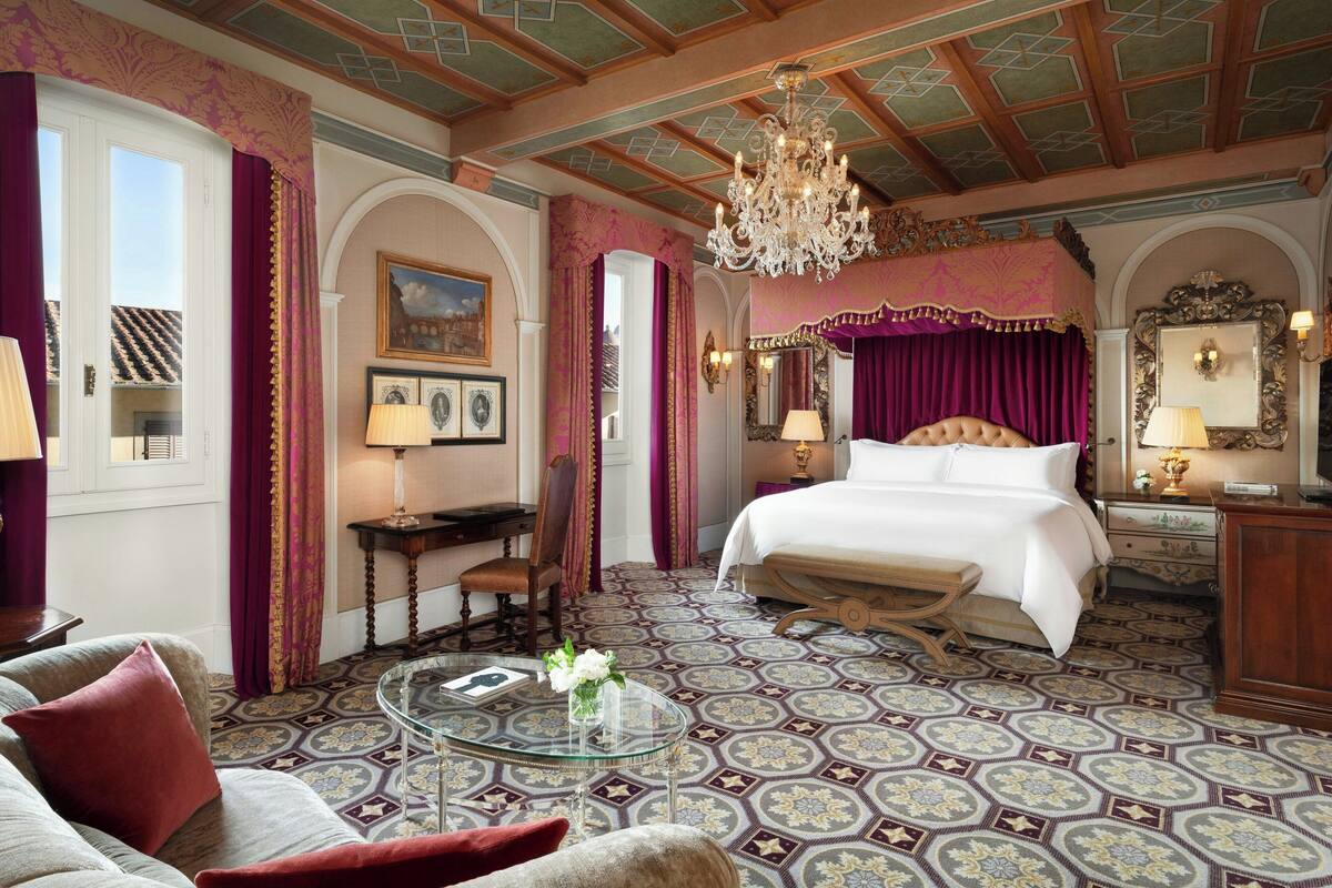 A luxurious hotel room with a large bed, a chandelier, a glass coffee table, and ornate floor tiles.