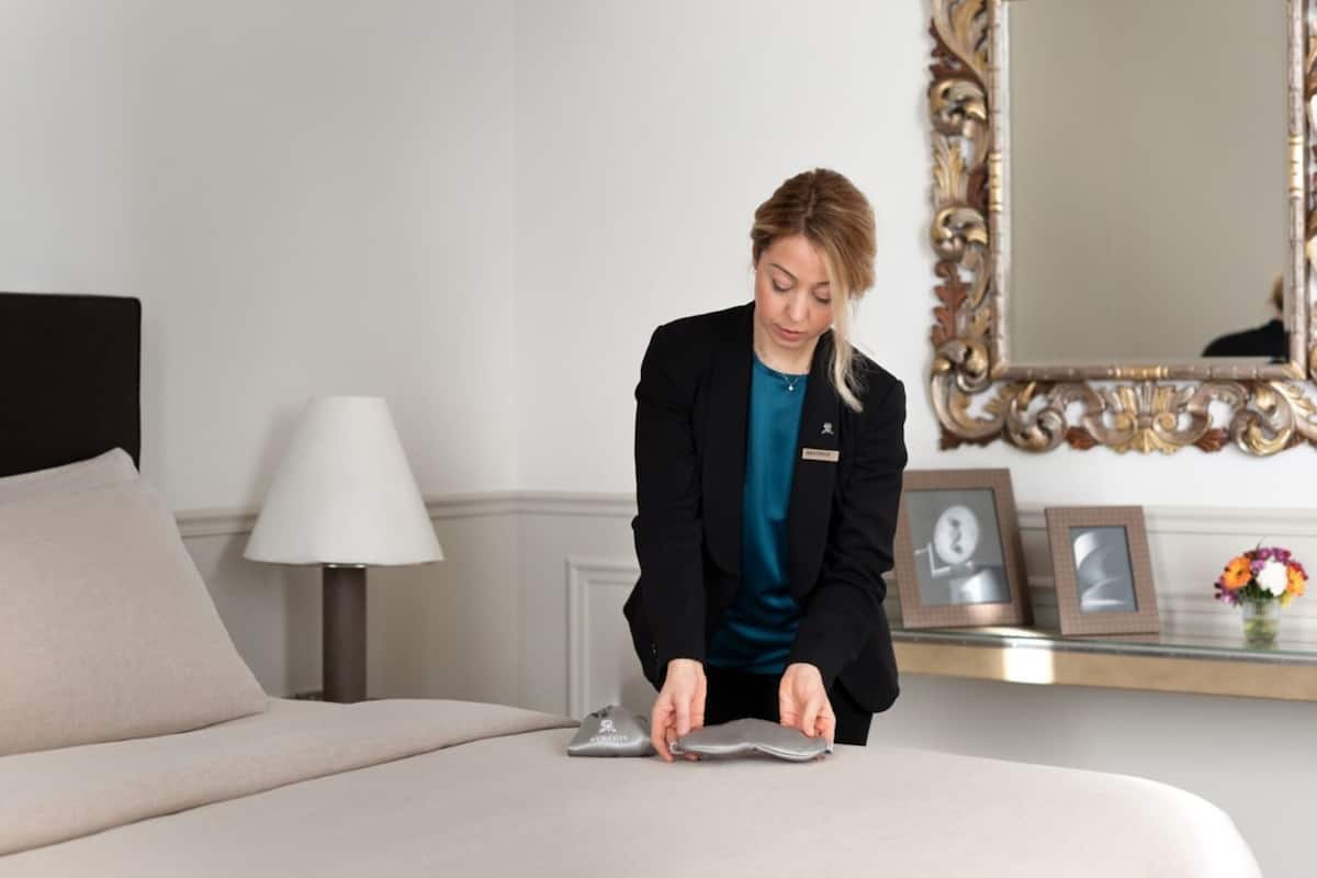 A hotel room with a bed, a lamp, a mirror, and a small table with framed pictures and a vase of flowers.