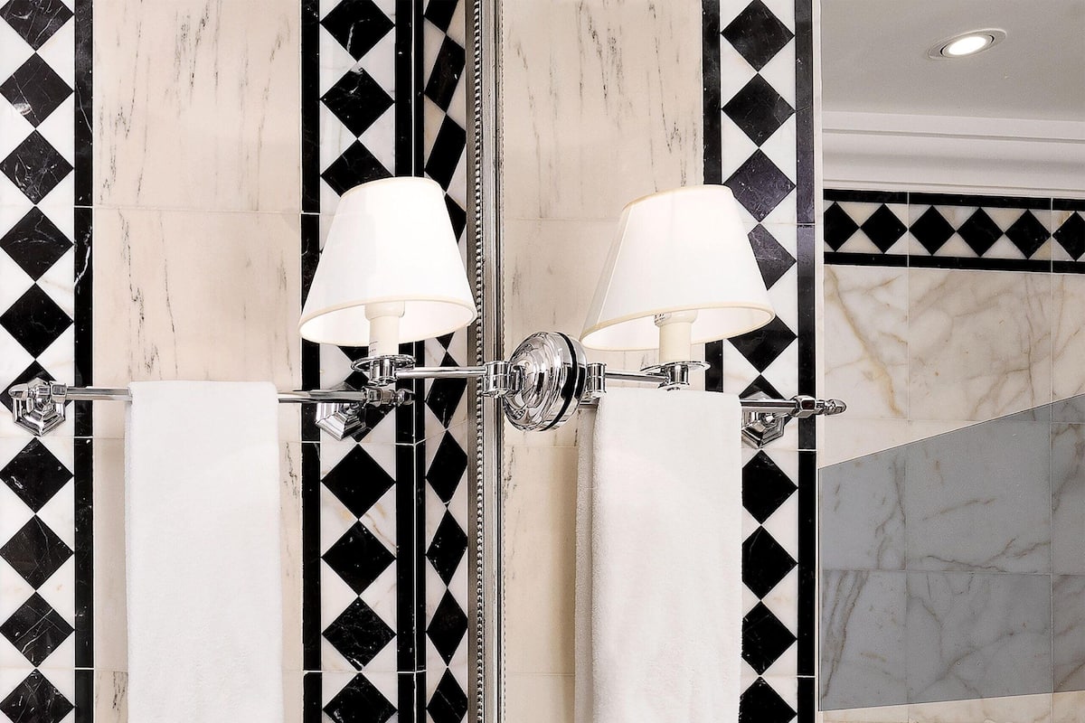 A bathroom with a black and white tiled wall, a large mirror, two wall-mounted lamps, a white sink, and a towel rack with two towels.