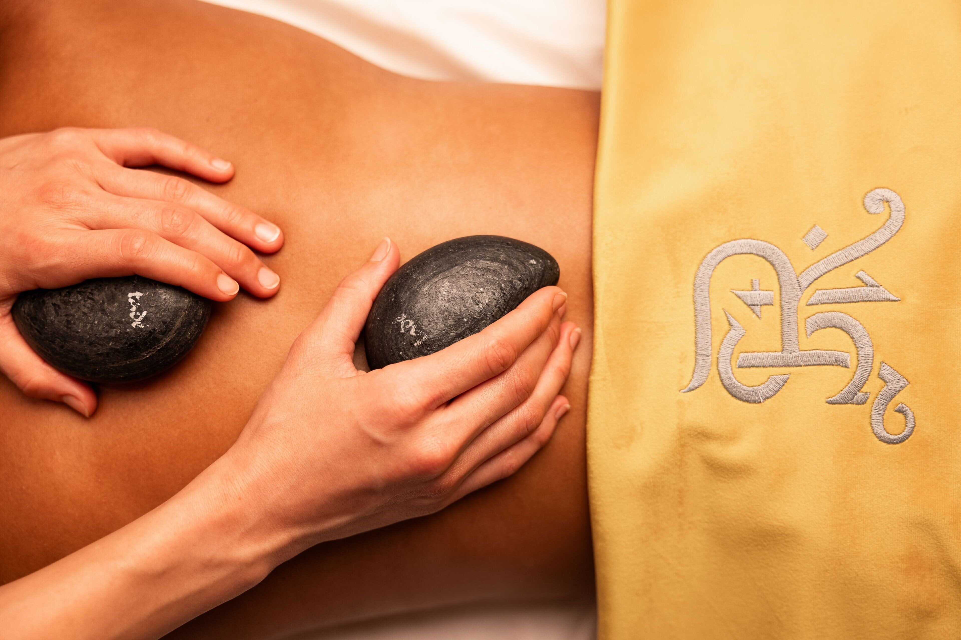 Gros plan d’un massage effectué à l’aide de deux pierres noires sur le dos d’une personne, avec une serviette jaune ornée de motifs brodés.