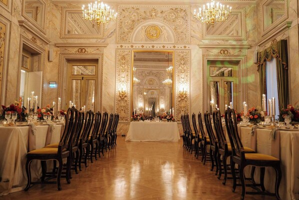 A grand banquet hall with ornate ceiling decorations, chandeliers, and a long table set for a formal event.