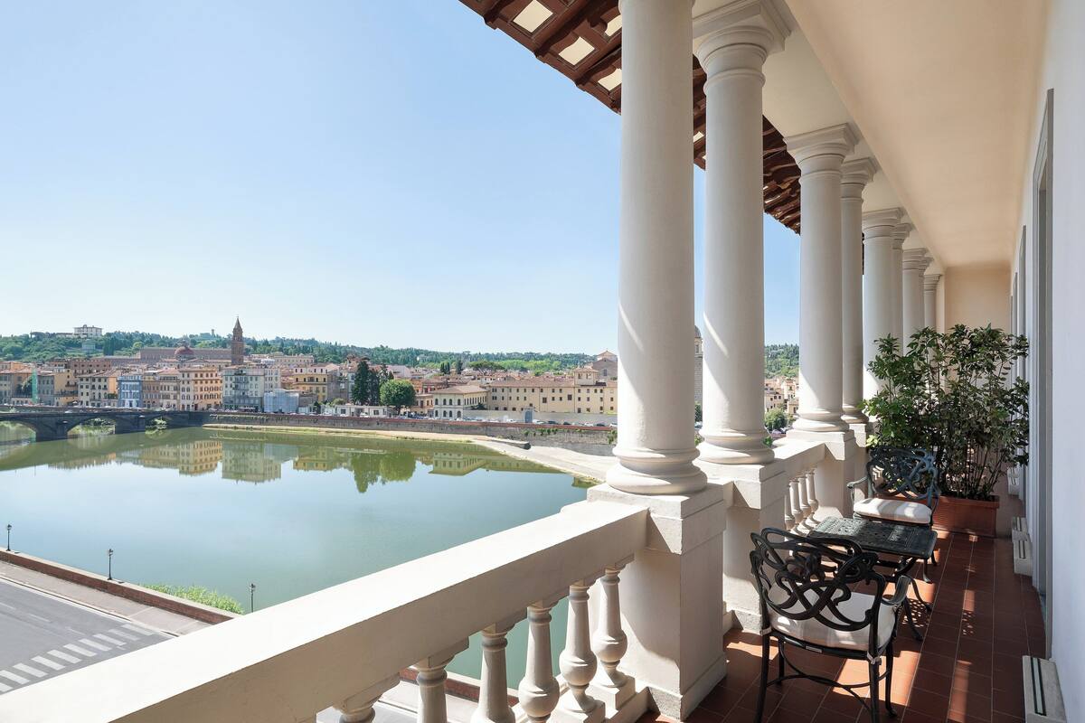A balcony with a view of a river and buildings, featuring a table and chairs.