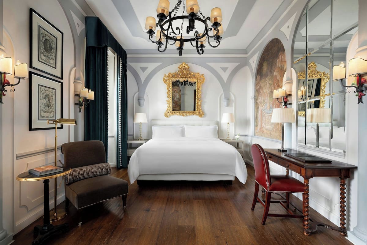 A hotel room with a large bed, a desk, a chair, a chandelier, framed artwork, and a mirror.
