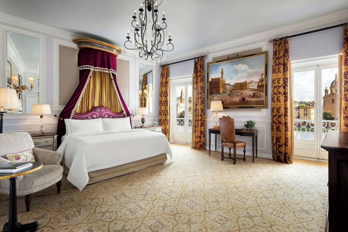 A luxurious bedroom with a four-poster bed, a chandelier, and a view of a cityscape through a large window.