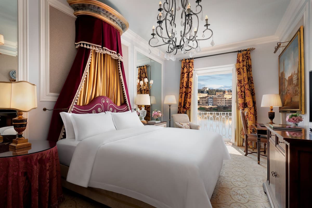 A luxurious hotel room with a large bed, ornate curtains, a chandelier, and a view of the city.