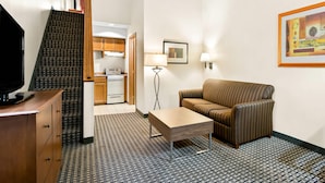 A compact living space with a sofa, a coffee table, a TV on a wooden cabinet, a patterned carpet, a framed artwork, and a kitchen in the background. - Quality Inn Mount Vernon (Mount Vernon)