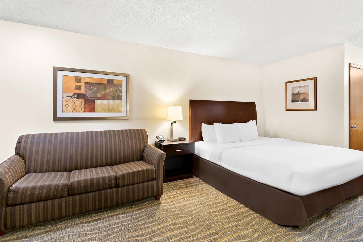 A hotel room with a striped sofa, a bed with white linens, a nightstand with a lamp, and framed artwork on the walls.