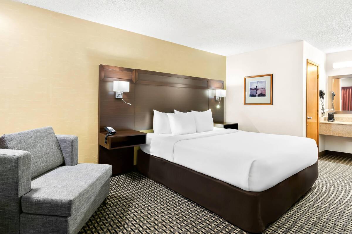 A hotel room with a large bed, a gray sofa, a wooden nightstand with a phone, and a framed picture on the wall.