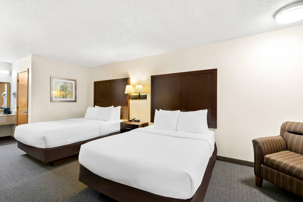A hotel room with two beds, a nightstand, a lamp, a chair, and a painting on the wall.