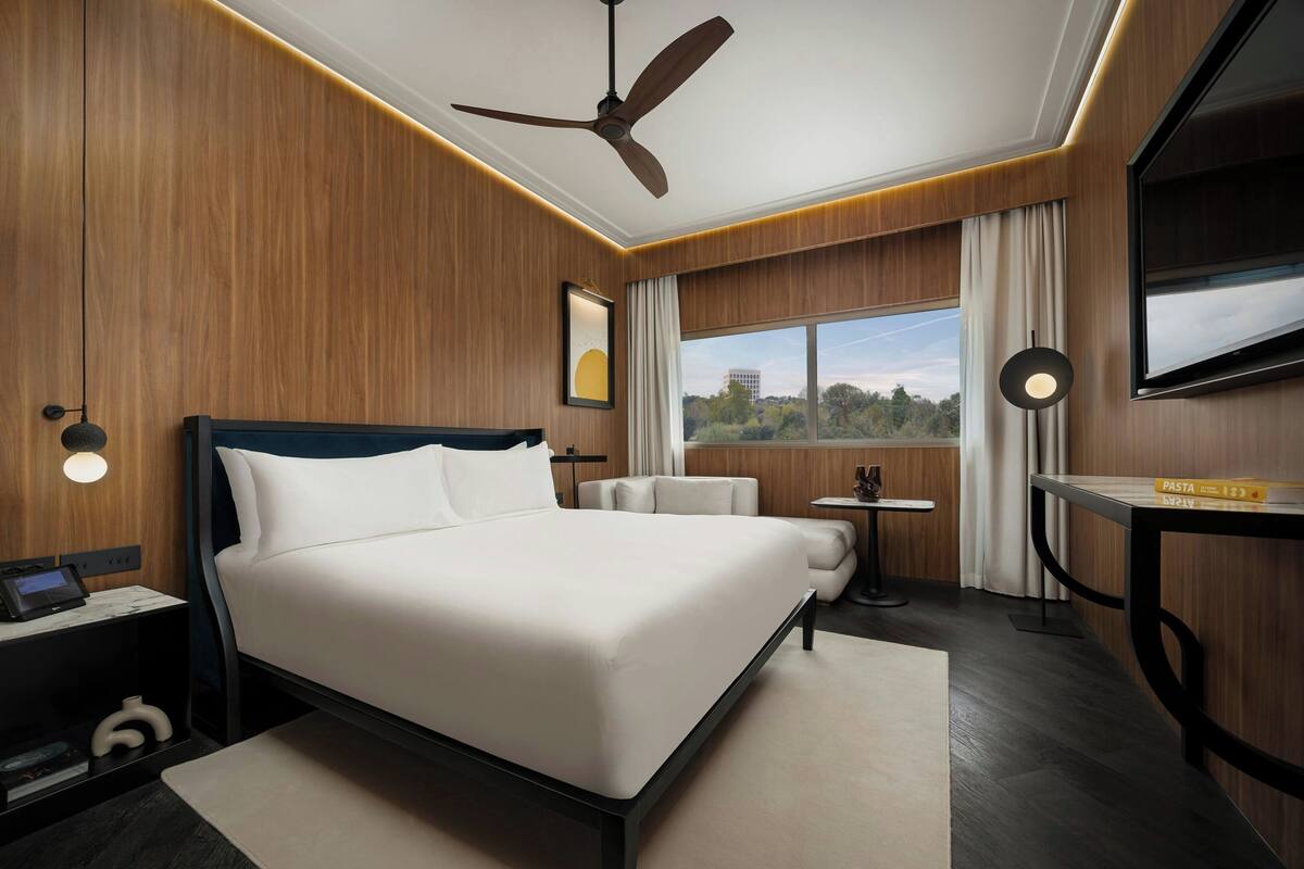 A modern hotel room with a large bed, a ceiling fan, a TV, a small table, and a window with a city view.