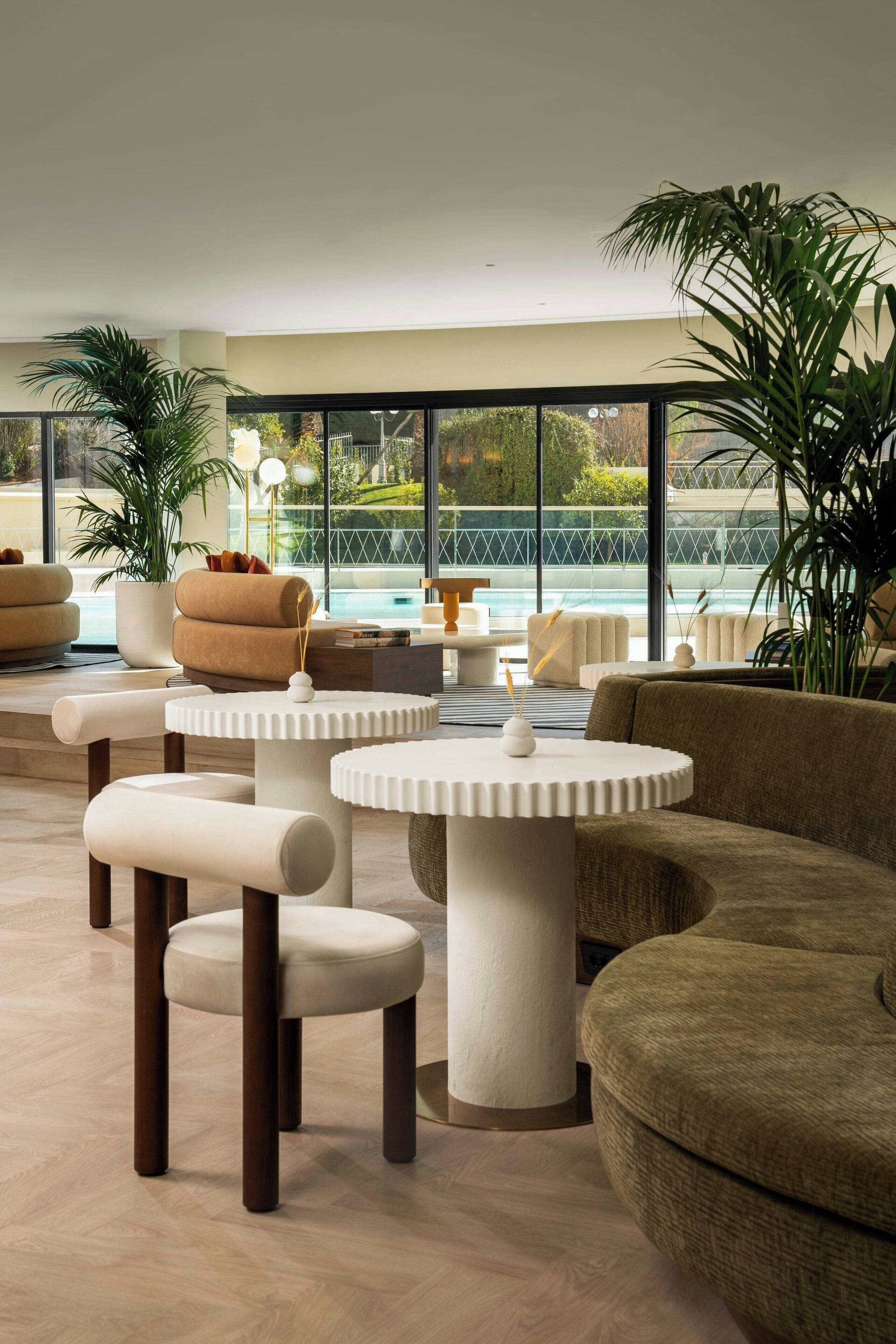 A modern lounge area with round tables and comfortable seating, large windows offering a view of a pool and greenery.