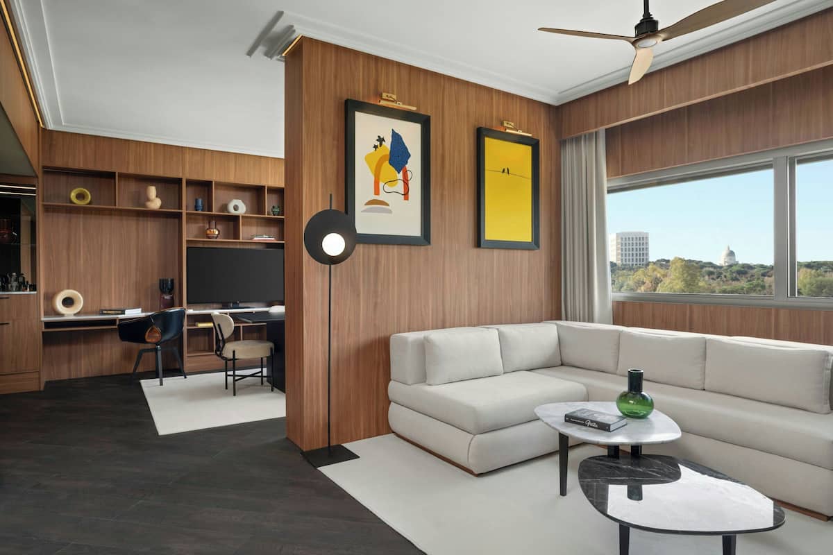 A modern living room with a sectional sofa, a coffee table, and a desk area with a chair. There are framed artworks on the wall and a view of the city through large windows.