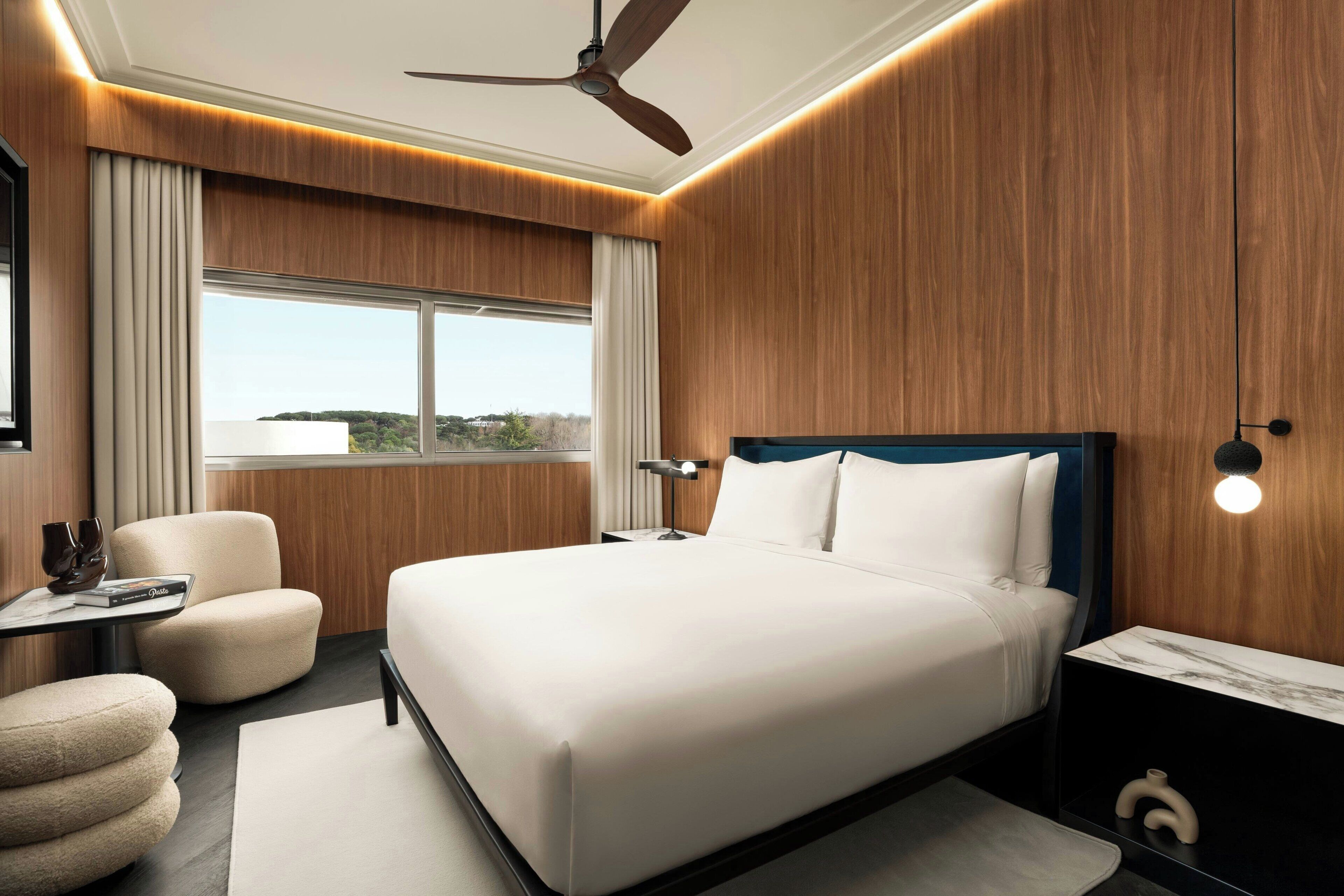 A modern hotel room with a large bed, a desk, a chair, and a view of the outdoors.