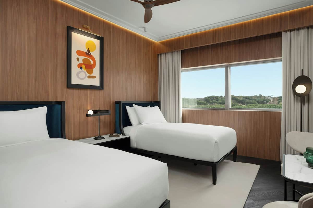 A hotel room with two beds, a large window with a view, a ceiling fan, and a framed picture on the wall.