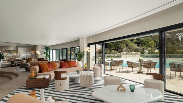 Un vaste espace de vie avec une grande fenêtre donnant sur une piscine et un jardin. On y trouve différents types de sièges, notamment des canapés et des fauteuils.