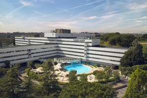 A modern building with a swimming pool, surrounded by greenery and a clear sky.