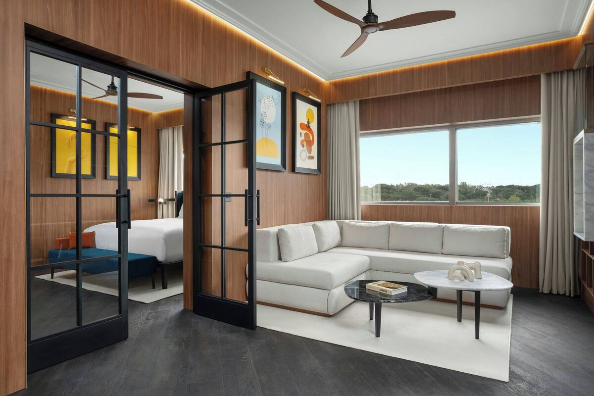 A modern hotel room with a large sectional sofa, a glass partition, a ceiling fan, and a large window offering a city view.