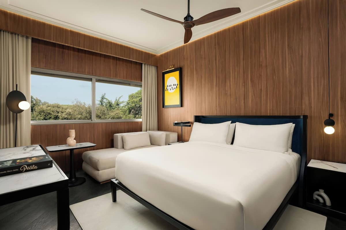 A modern hotel room with a large bed, a small sofa, a side table, and a window with a view of greenery.
