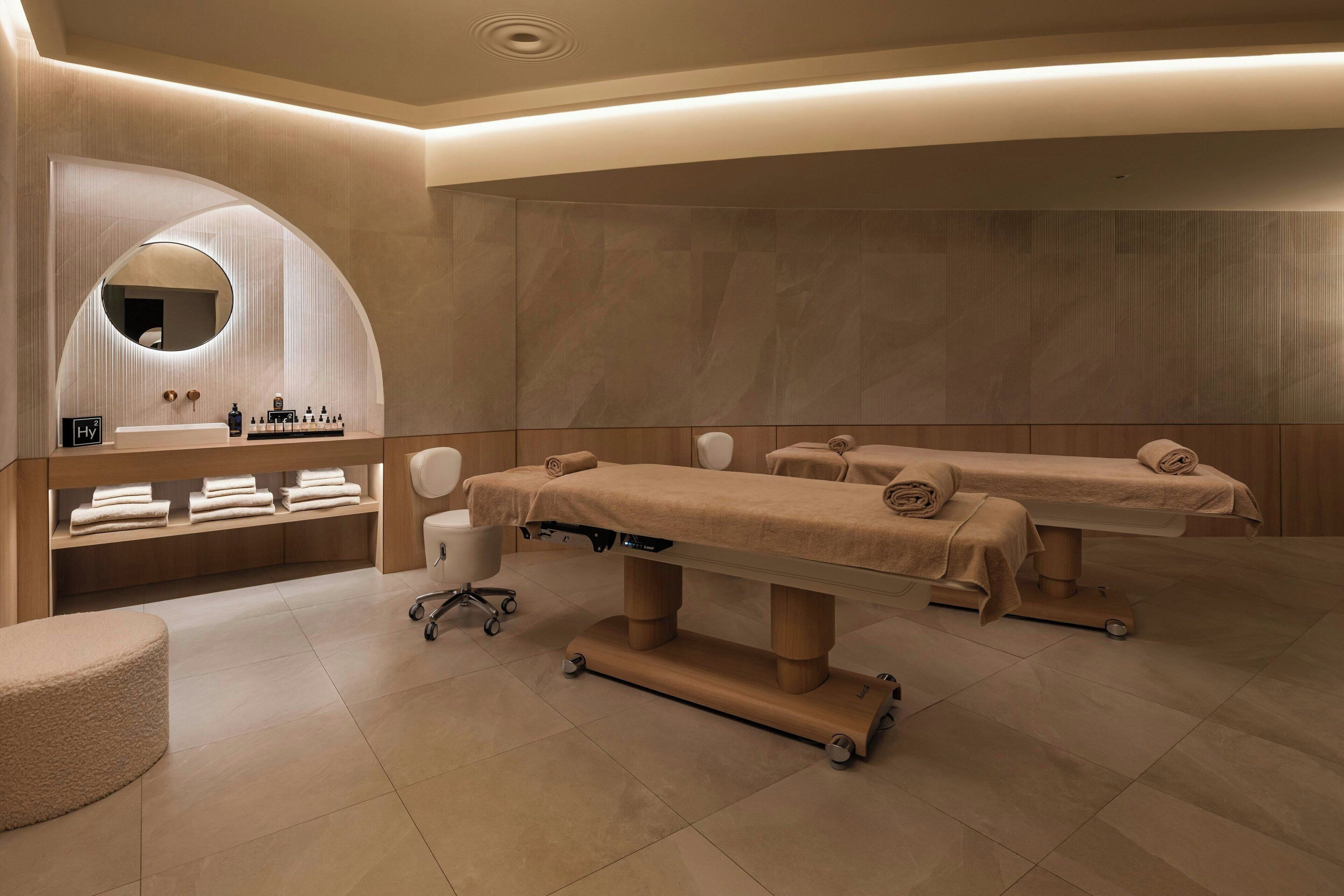 A modern spa room with two treatment tables, a massage chair, and a vanity with a mirror and toiletries.
