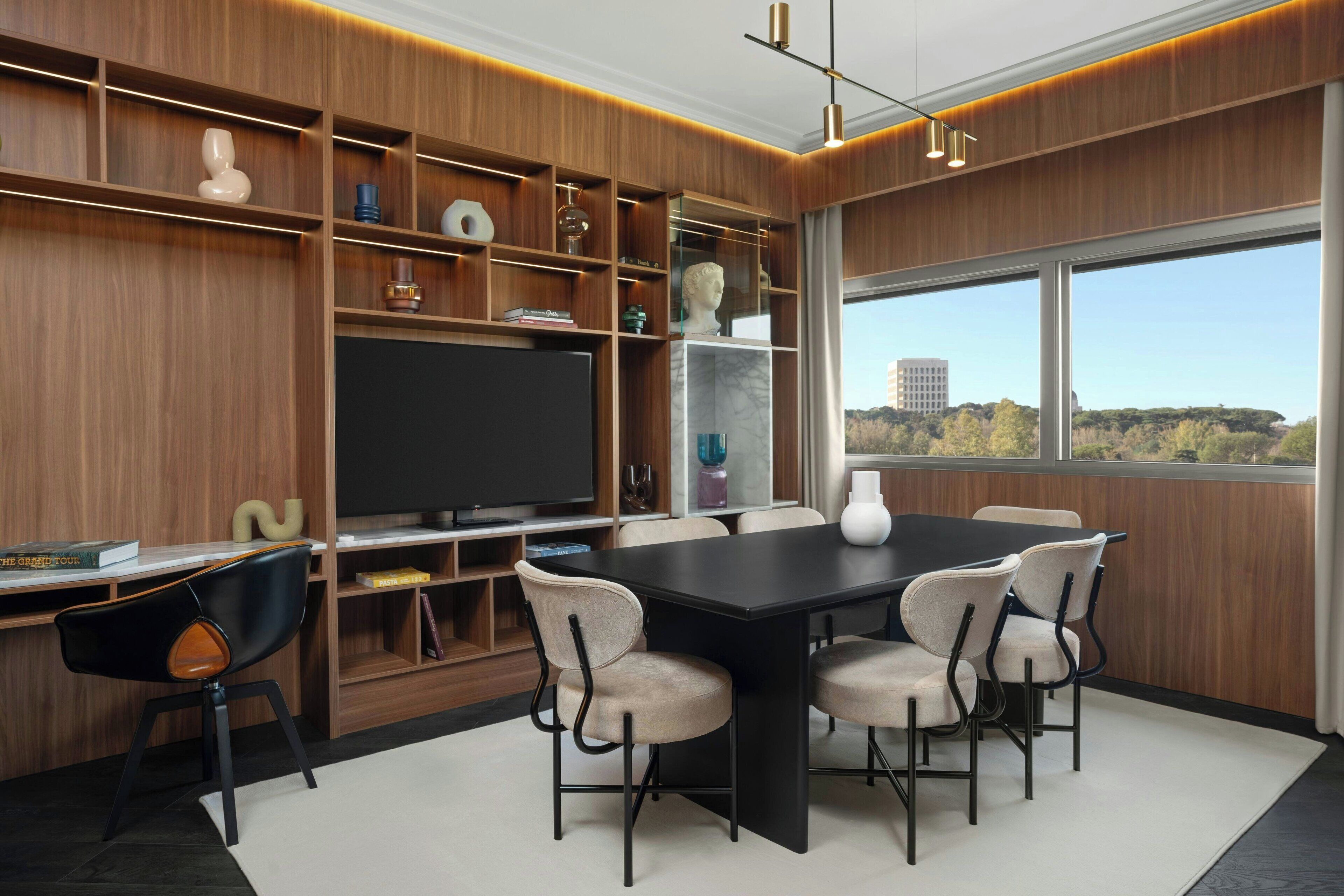 A modern dining area with a large table and chairs, a television, and a view of a cityscape through large windows.