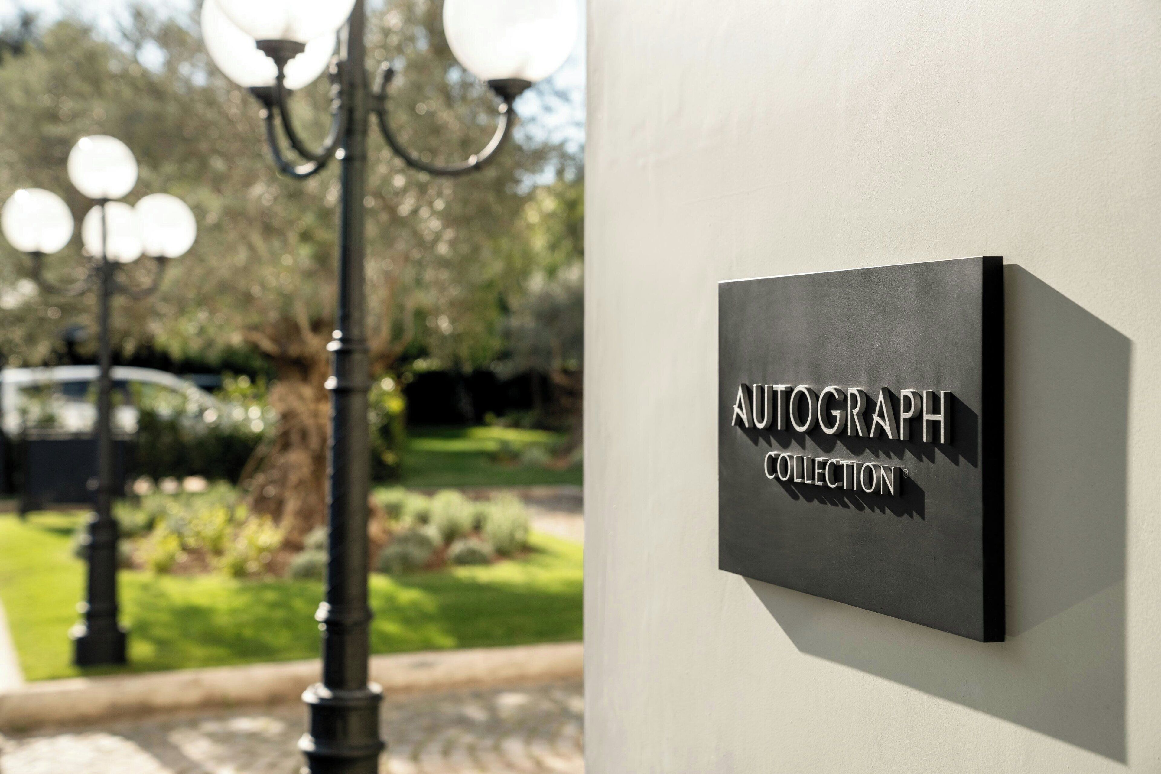 Hyvin hoidetulla ulkoalueella, jossa on valaisinpylväs ja viheralueita, on seinälle kiinnitetty kyltti, jossa lukee: ”AUTOGRAPH COLLECTION”.