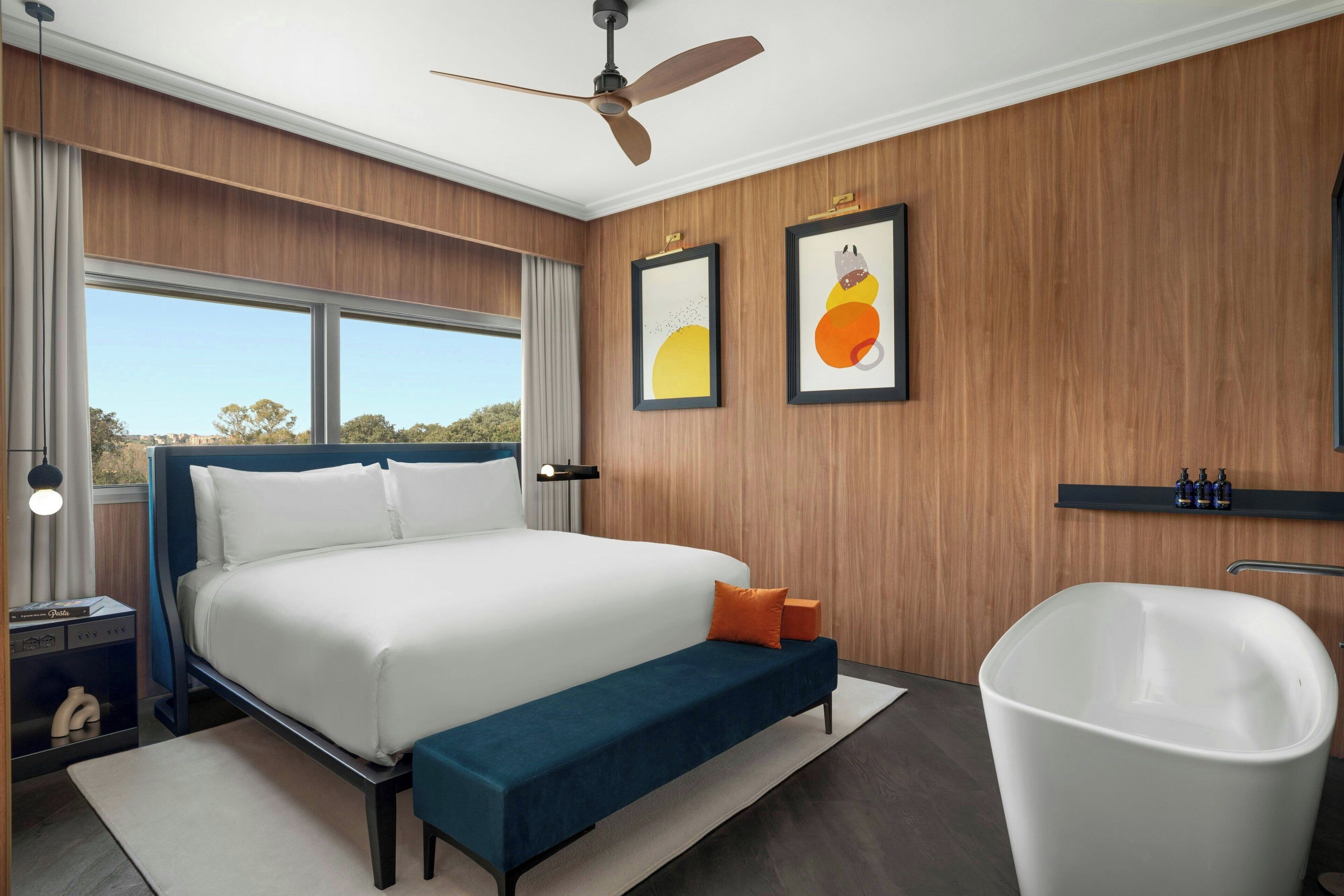 A modern bedroom with a large bed, a bench, a bathtub, and artwork on the wall.