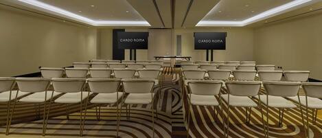 Un salón de conferencias con filas de sillas blancas, una mesa central y dos pantallas mostrando