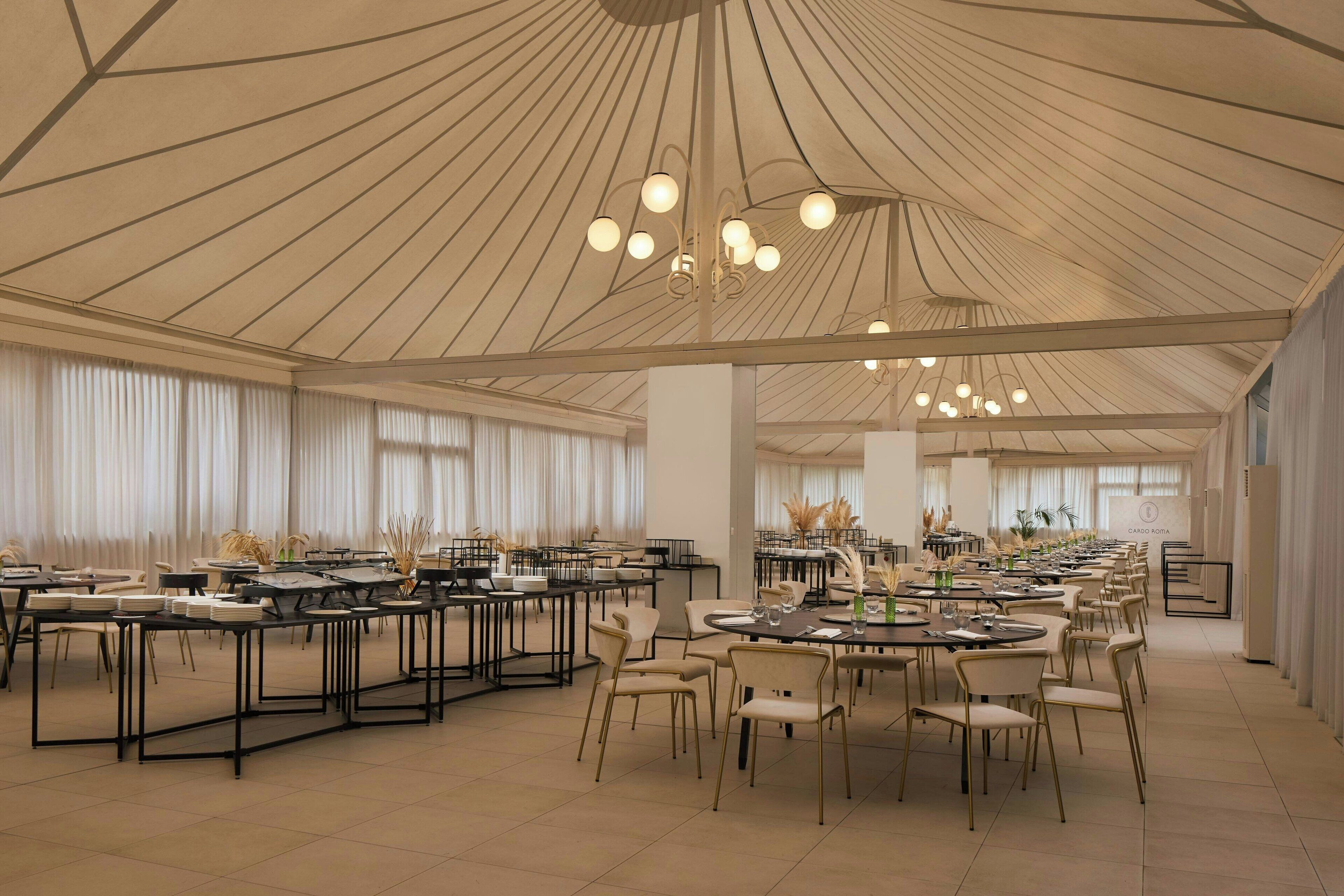 Une salle de banquet spacieuse, dotée de nombreuses tables et chaises rondes, d’un plafond haut orné d’un motif radial original et de grandes fenêtres avec des rideaux.