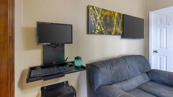 Quarto com um sofá cinzento, uma secretária com um computador, um monitor e um objeto decorativo na parede.