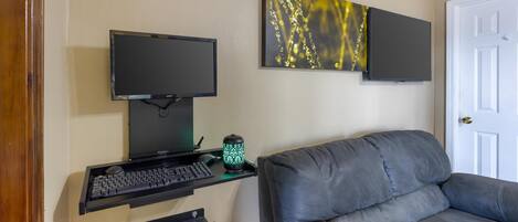 Una habitación con un sofá gris, un escritorio con computadora, un monitor y un adorno en la pared.