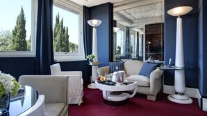 A luxurious room with a red carpet, a round coffee table, and a sofa. There are two tall lamps and a window with curtains. - Hotel Lord Byron (Rome)