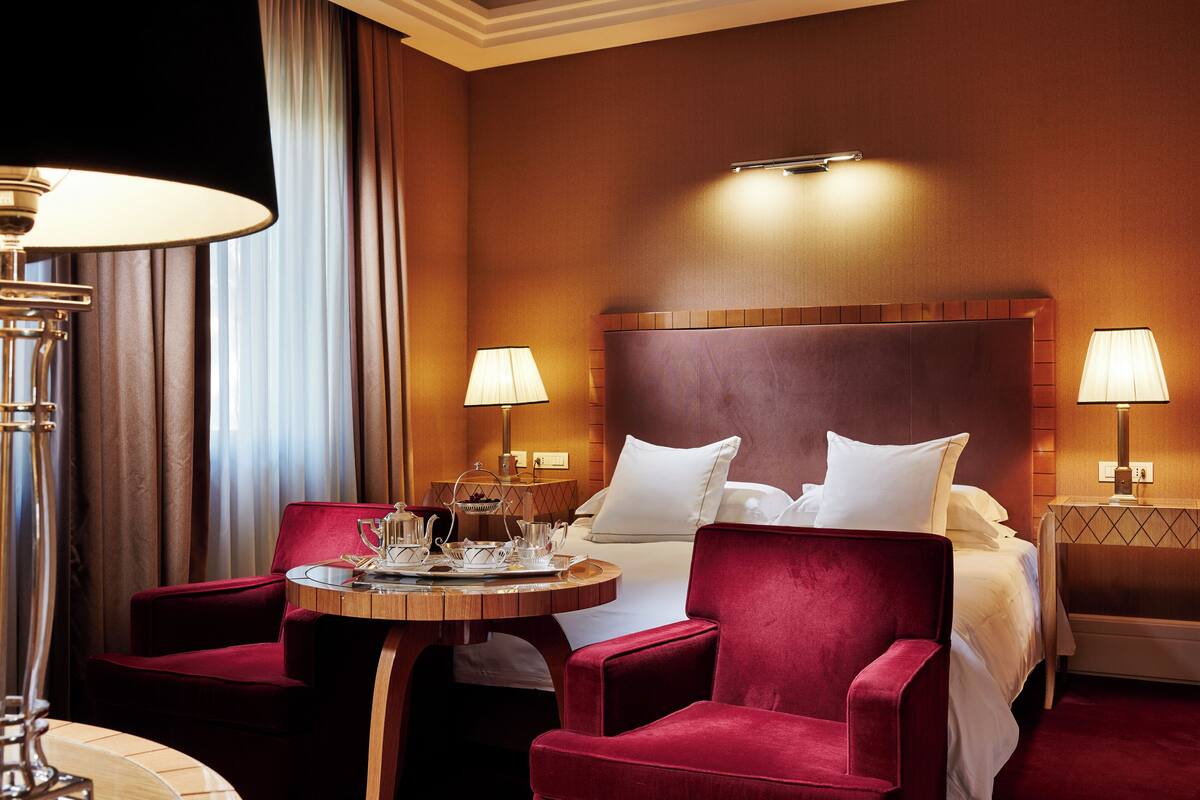 Hotellihuone, jossa on sänky, kaksi punaista tuolia, pyöreä pöytä tarjottimella ja kaksi yöpöytävalaisinta.