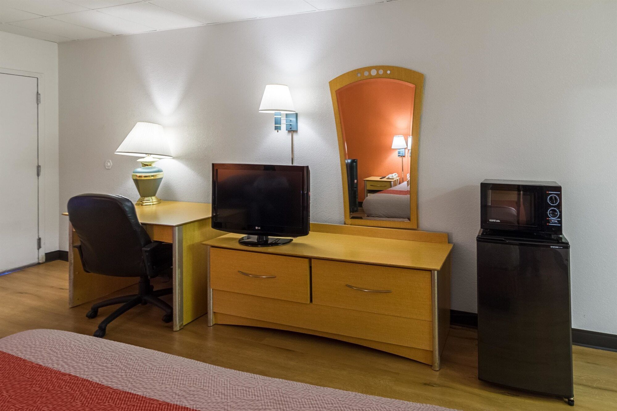 A hotel room with a wooden desk, a computer monitor, a flat-screen TV, a microwave, a chair, and a bedside table with a lamp.