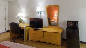 A hotel room with a wooden desk, a computer monitor, a flat-screen TV, a microwave, a chair, and a bedside table with a lamp.