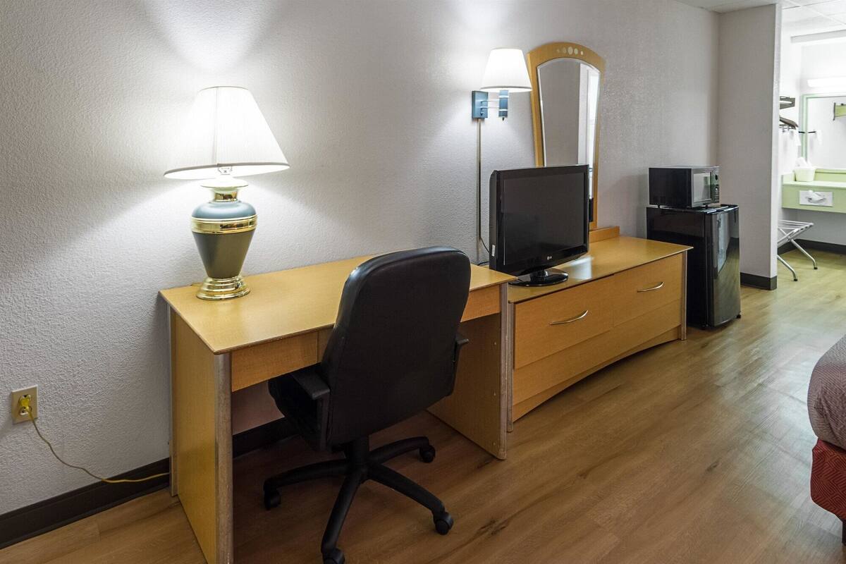 A hotel room with a wooden desk, a black office chair, a flat-screen TV, a lamp, and a mirror.