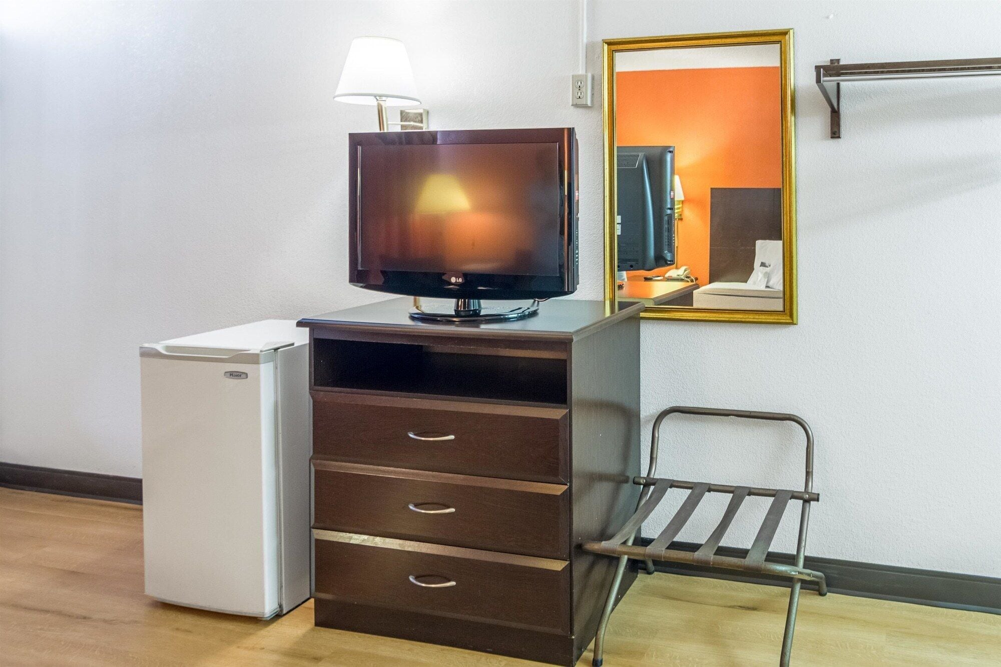 A compact room with a flat-screen TV, a small refrigerator, a wooden cabinet with drawers, a metal chair, and a mirror on the wall.